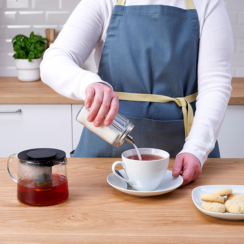 Una persona con un delantal vierte té en una taza de un recipiente de vidrio. Una tetera con infusor y galletas están sobre la mesa.
