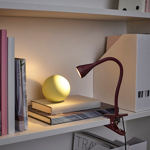 Una lámpara de abrazadera NÄVLINGE de color burdeos en un estante, iluminando una esfera amarilla sobre libros. Delgado y ajustable, contrasta con libros y cajas más grandes.