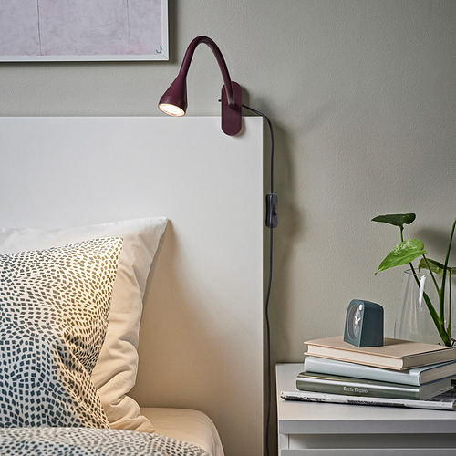 Lámpara de pared granate NÄVLINGE cerca de la cama. Diseño delgado con brazo ajustable, junto a una pila de libros y plantas.