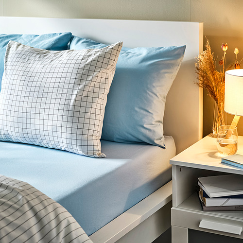 Cama azul con almohada de cuadros, almohada a rayas, lámpara, flores secas, libros.