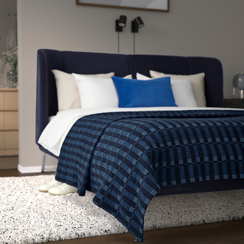 Dormitorio moderno con cama grande. Cabecero azul oscuro y manta a cuadros. Alfombra blanca, zapatillas blancas. Entorno agradable y acogedor.