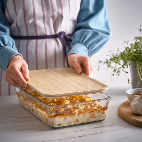 Una persona coloca una tapa de bambú en una fuente de vidrio para hornear con lasaña, demostrando el almacenamiento y la protección de los alimentos.