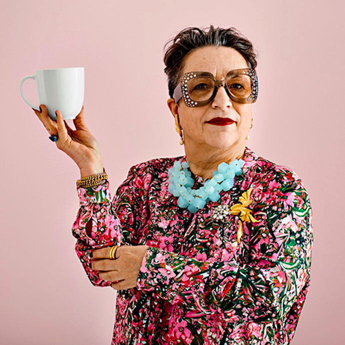 Mujer con estilo, gafas grandes y collar azul, levanta una taza blanca sobre fondo rosa.  