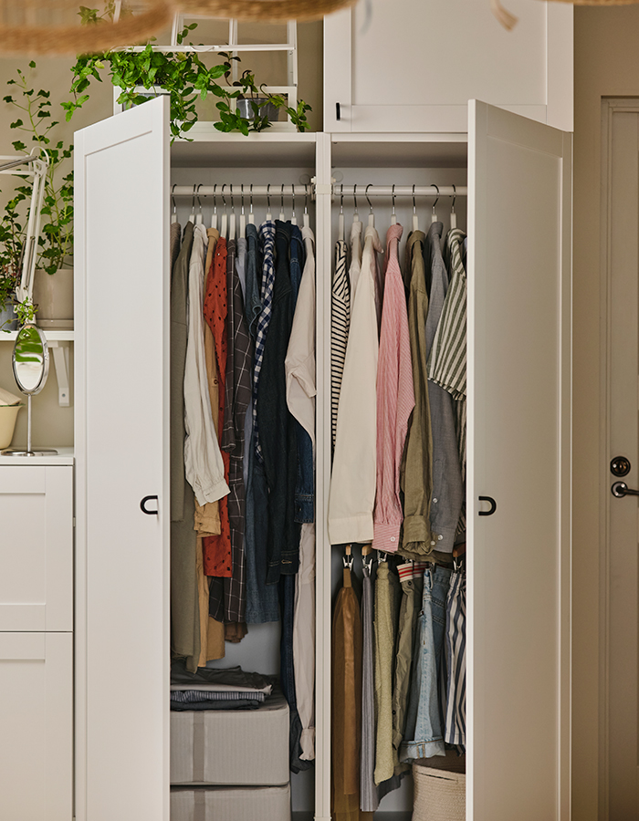 Open white wardrobe with color-coordinated hanging clothes, storage boxes at the bottom, and decorative plants on top.