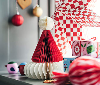 Decoración navideña de papel plegado en forma de gorro de Santa rojo y blanco sobre una cómoda. Fondo con cuadros y adornos colgantes.