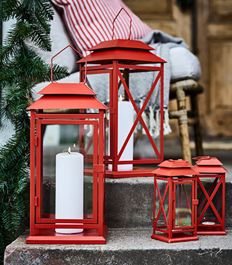 Faroles rojos de metal con velas blancas grandes en una escalera de piedra, junto a una guirnalda verde y un cojín a rayas.