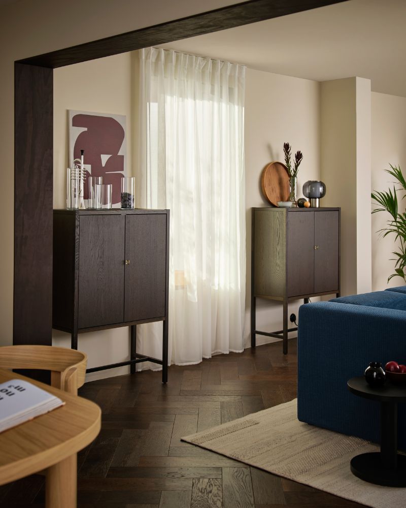 A living room with two dark-brown STOCKHOLM 2025 cabinets against a light wall and a window covered with sheer curtains.