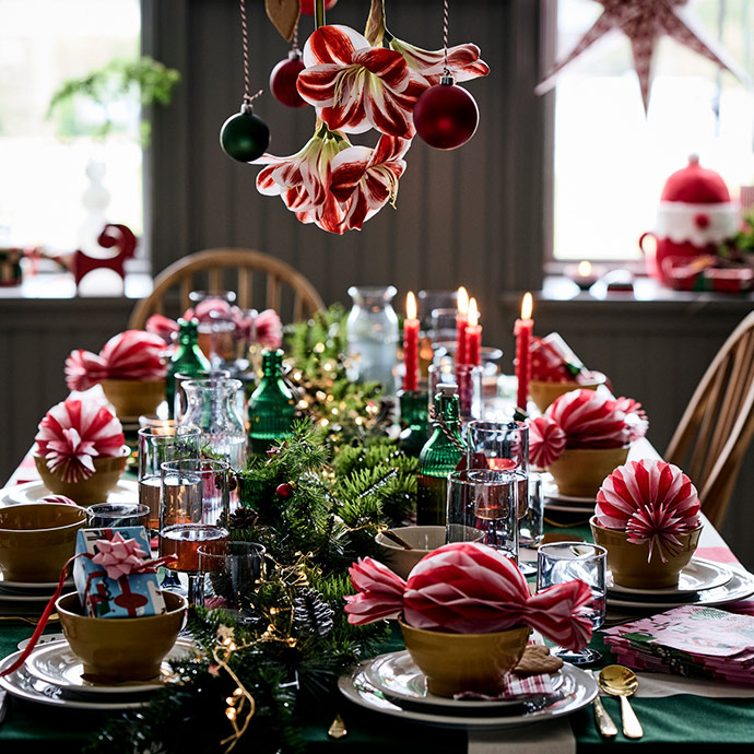 Mesa de comedor decorada para Navidad con mantel verde, centro de abeto, velas rojas, copas y servilletas de papel que parecen caramelos.