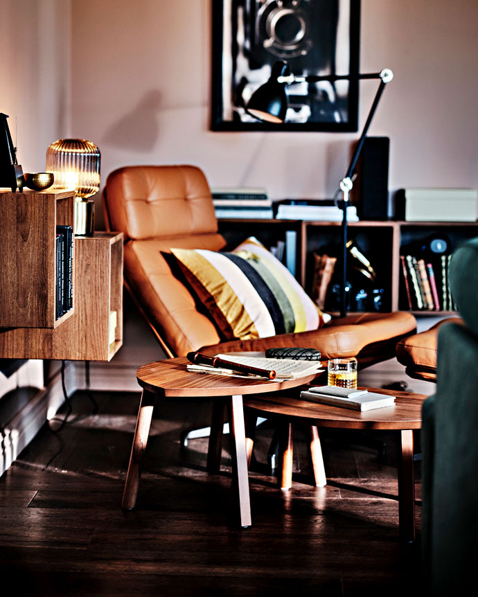 Brown leather armchair in a cozy living room with wooden nesting tables, low bookshelves, a black floor lamp, and warm sunset lighting.