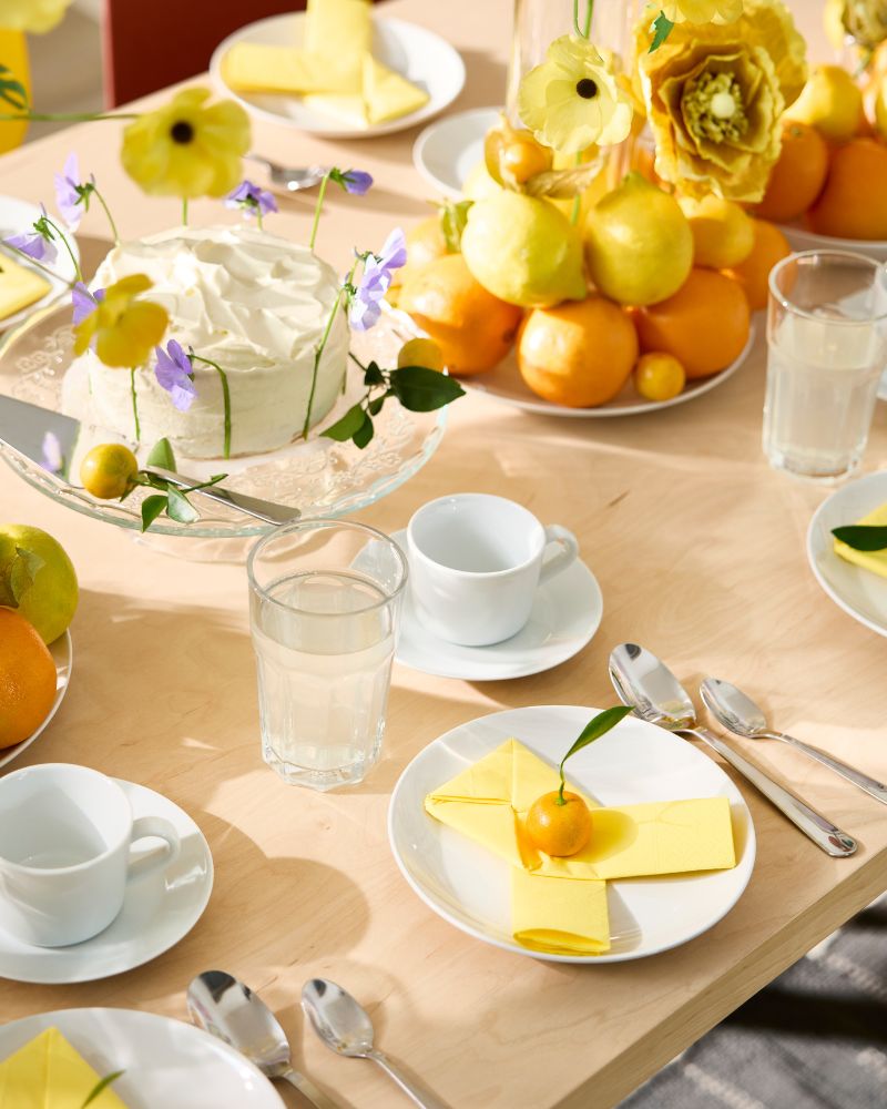 A birthday table setting has white plates with yellow paper napkins, POKAL glasses and plates with oranges and lemons.