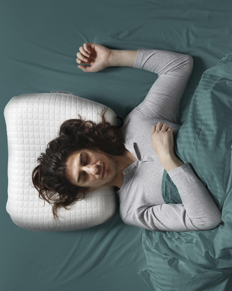 Vista aérea de una persona durmiendo boca arriba con el brazo levantado y la cabeza sobre una almohada ergonómica KLUBBSPORRE.