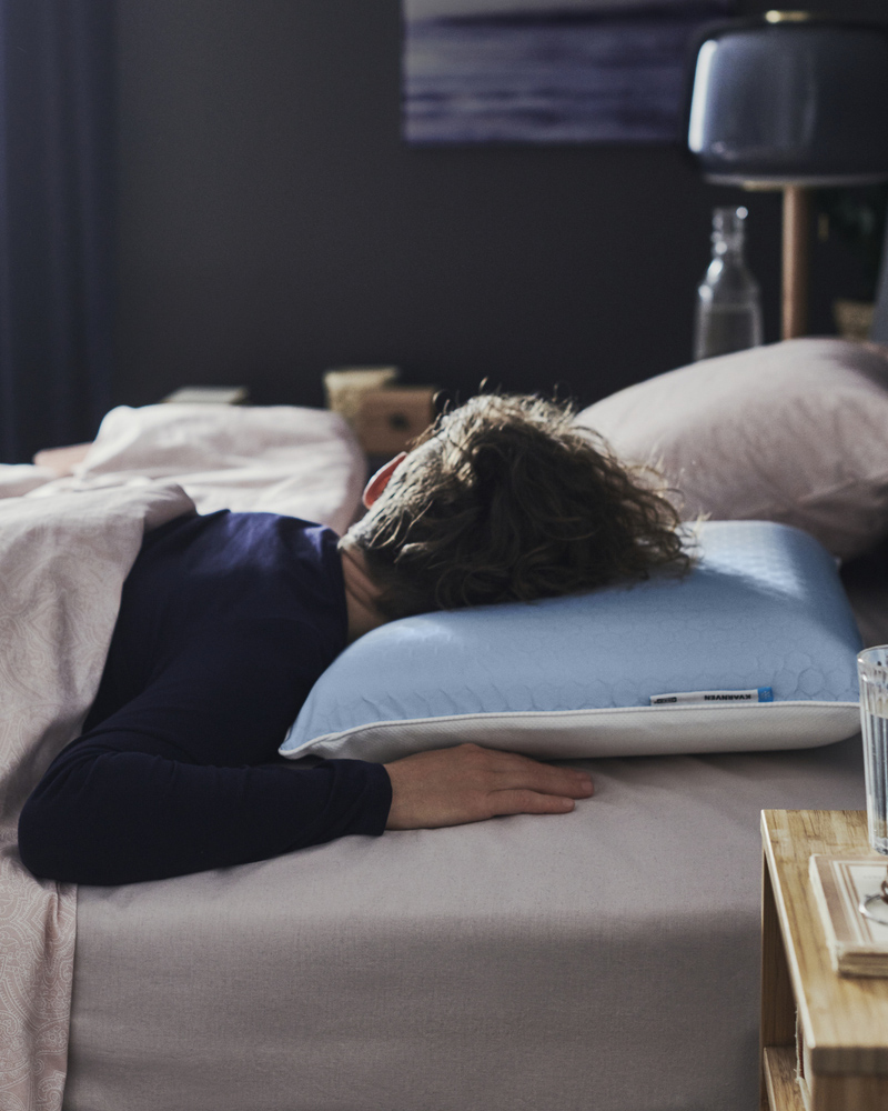 En un dormitorio oscuro, una persona en pijama azul duerme boca abajo en la cama con una almohada ergonómica KVARNVEN.