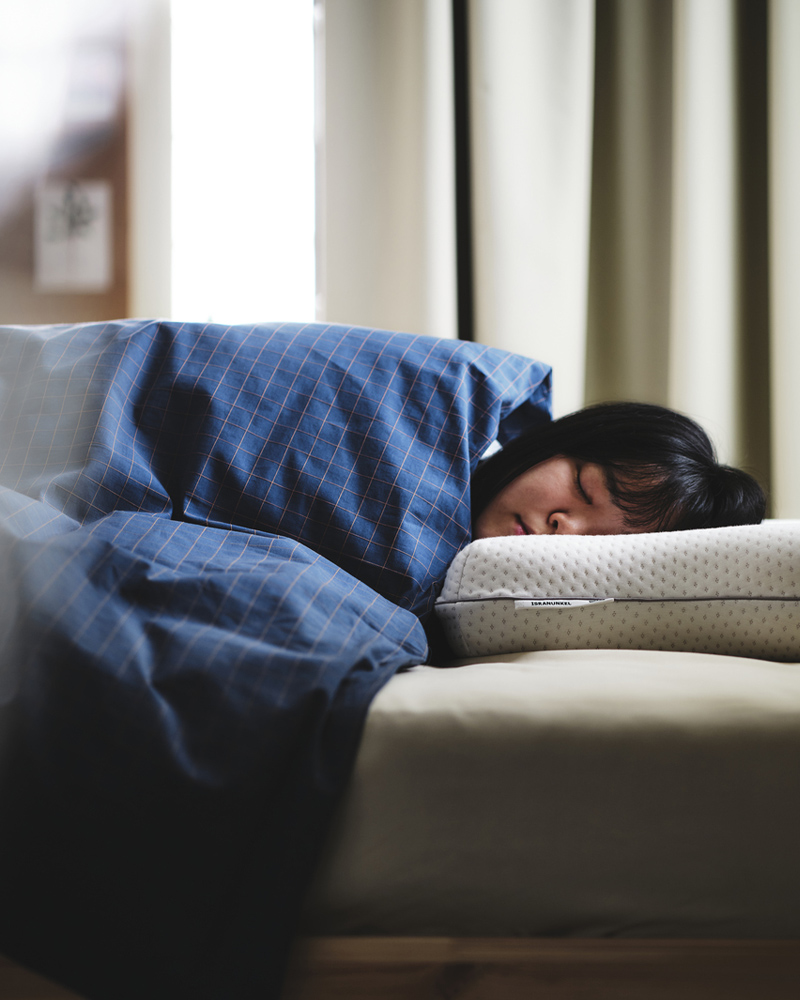 Una persona durmiendo bajo sábanas DOFTAKLEJA de color azul y marrón con la cabeza sobre una almohada ergonómica multiposición ISRANUNKEL.