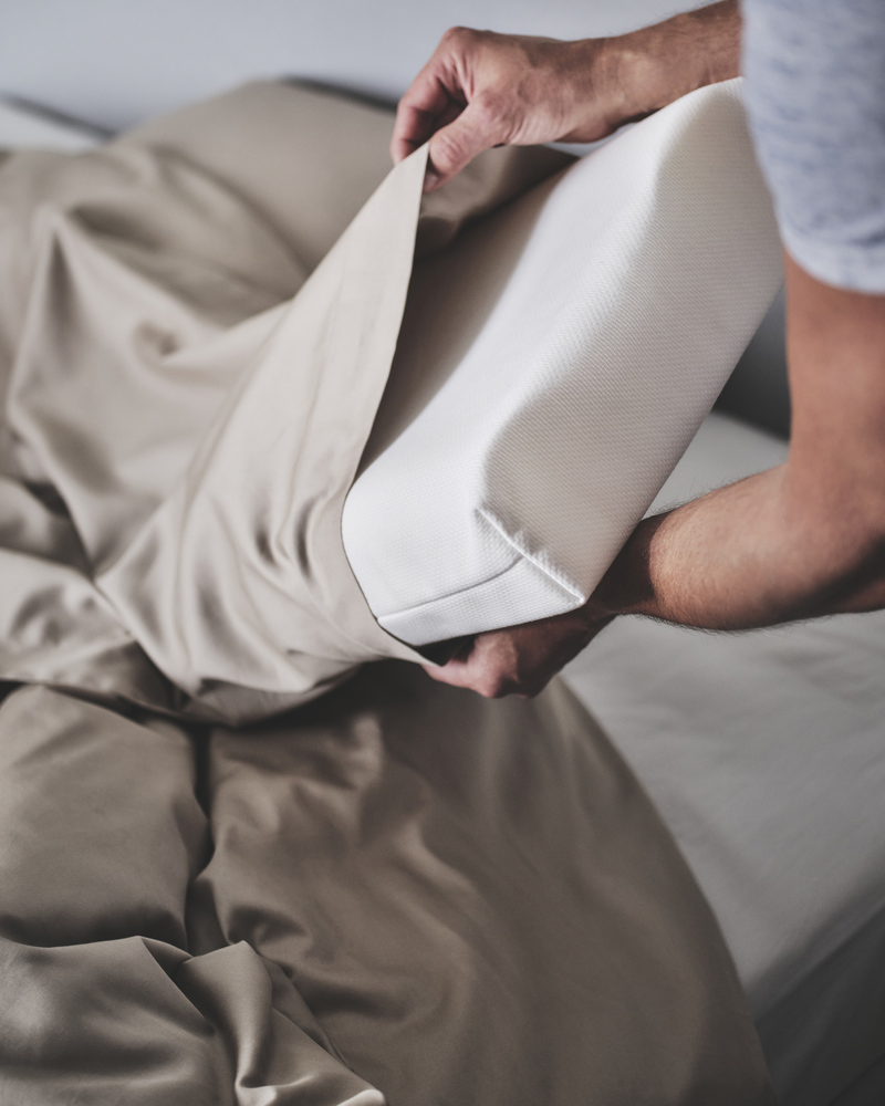 Dos manos sosteniendo la almohada ergonómica PAPEGOJBUSKE blanca para dormir de lado o boca arriba y ropa de cama de satén beige relajante.