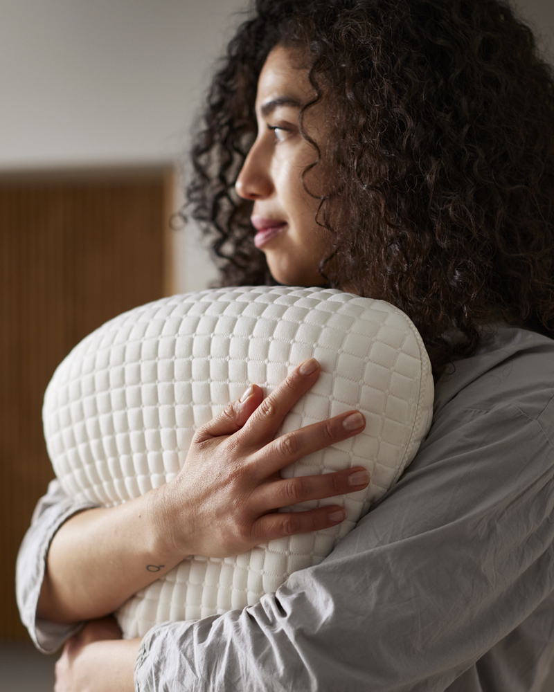 Una persona sosteniendo una almohada ergonómica KLUBBSPORRE para quienes duermen de lado y boca arriba contra su pecho.