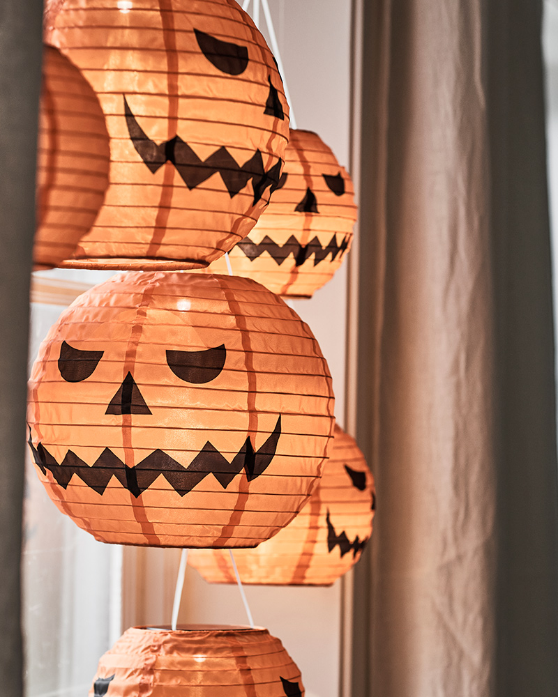 Some KUSTFYR pendant lamps resembling carved pumpkins are hanging in front of a window, next to a curtain