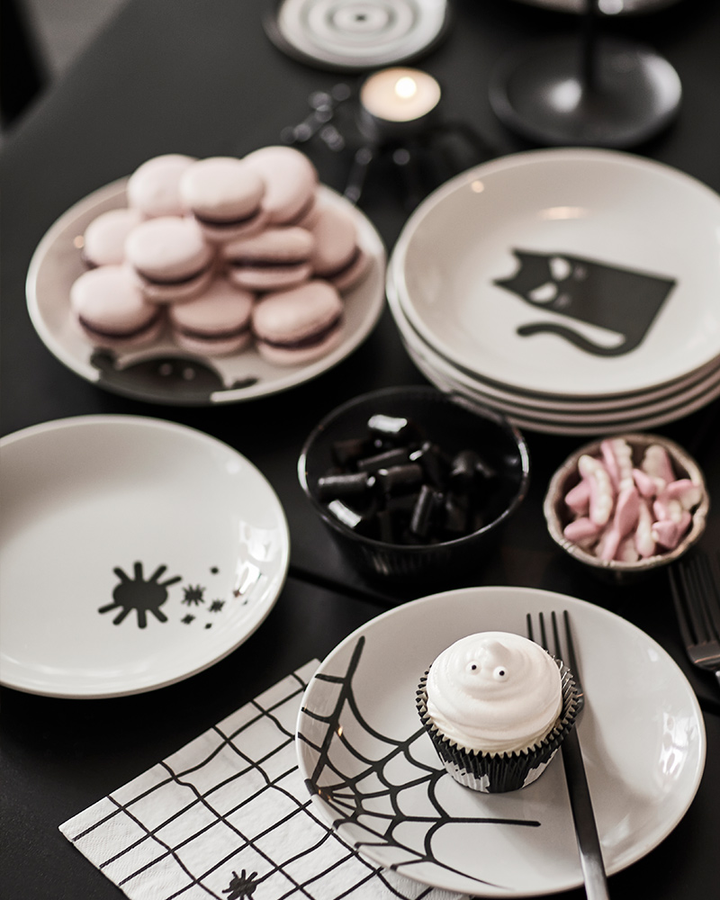 Several KUSTFYR side plates with Halloweenthemed decor are placed on a black table along with treats and decorations.