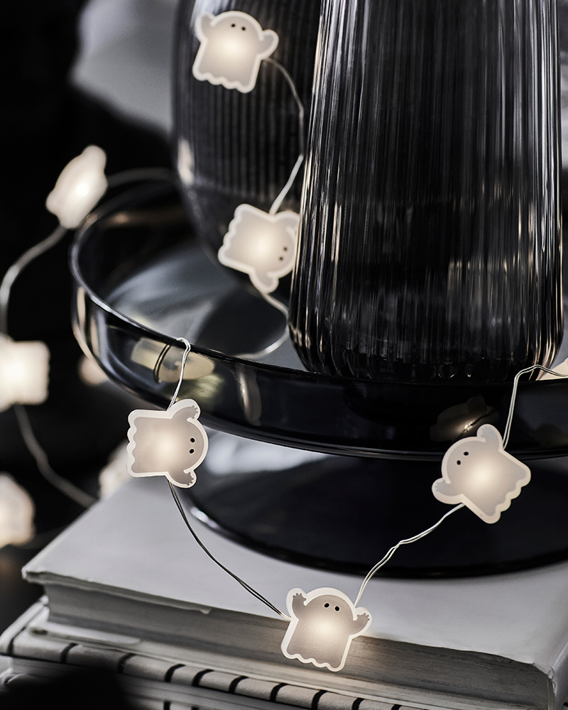 A KUSTFYR light chain featuring several small ghosts is wrapped around a black decoration and placed on top of some books.