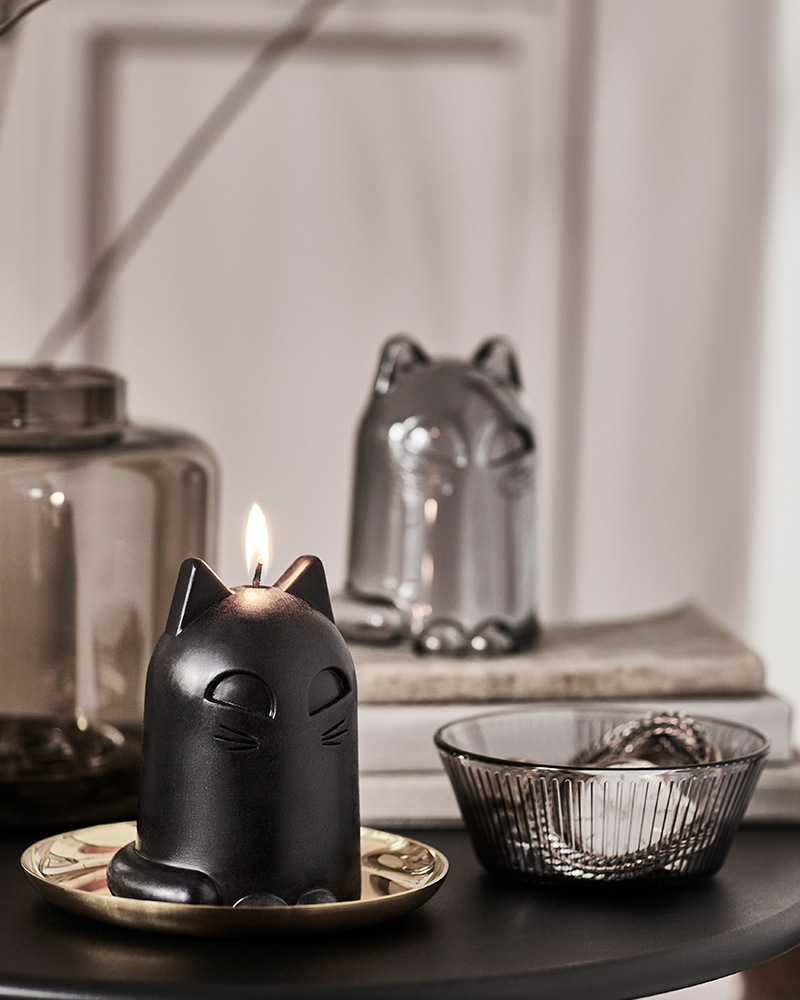 A KUSTFYR unscented candle and a KUSTFYR glass dome, both shaped like cats, are placed on a black table next to a bowl.