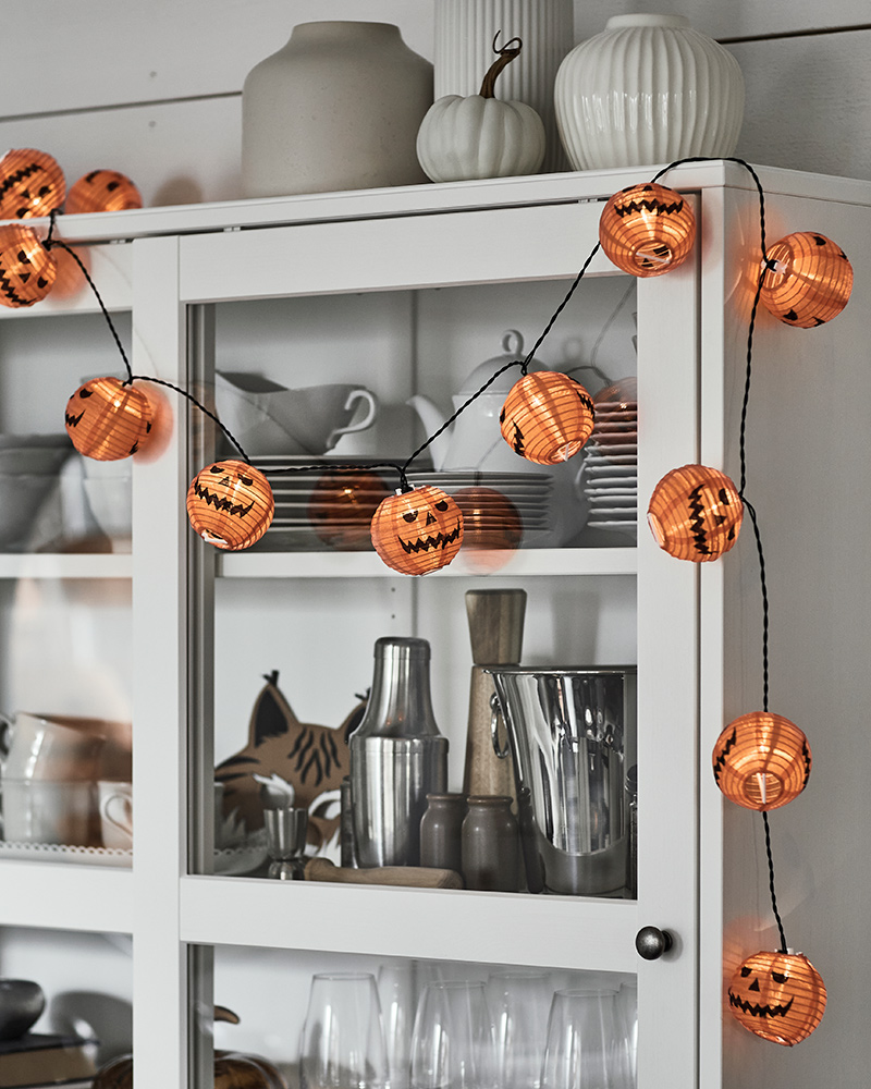 A KUSTFYR light chain featuring several small carved pumpkins is placed on top of a white cabinet with glass doors.
