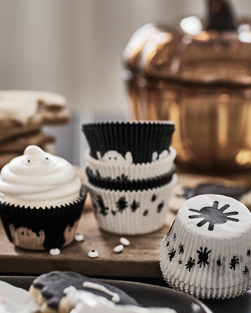 Some KUSTFYR baking cups decorated with ghosts and spiders are on a table next to a cupcake, some cookies, and a glass bowl.
