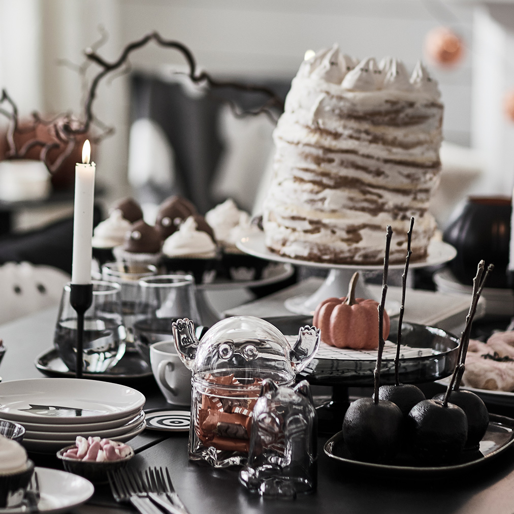 A dark table with KUSTFYR side tables, several treats, a KUSTFYR bowl with lid shaped like a ghost and some lit candles.