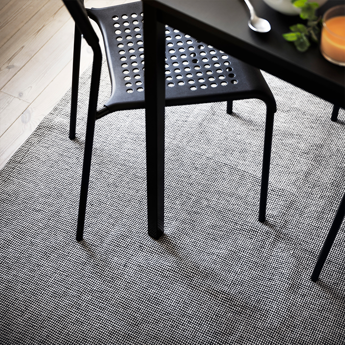 Alfombra bicolor de patrón geométrico bajo una mesa y sillas de comedor negras con patas finas, sobre un suelo de madera clara, creando un ambiente moderno y funcional.