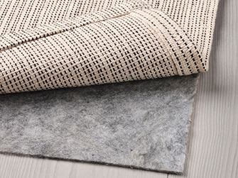 Detalle de la parte trasera de una alfombra de tejido natural, mostrando su textura y el reverso antideslizante, superpuesta sobre una base de fieltro gris para mayor confort y estabilidad.