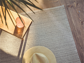 Alfombra de tejido plano en tonos neutros con una mesa auxiliar de madera y un sombrero de paja, bajo la luz del sol que entra por una ventana, creando sombras suaves.