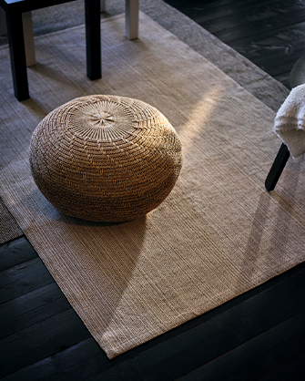 Alfombra de fibra natural en tono beige claro con un pouf de mimbre texturizado en el centro, y muebles con patas oscuras alrededor, en un espacio con suelo de madera oscura y luz natural.