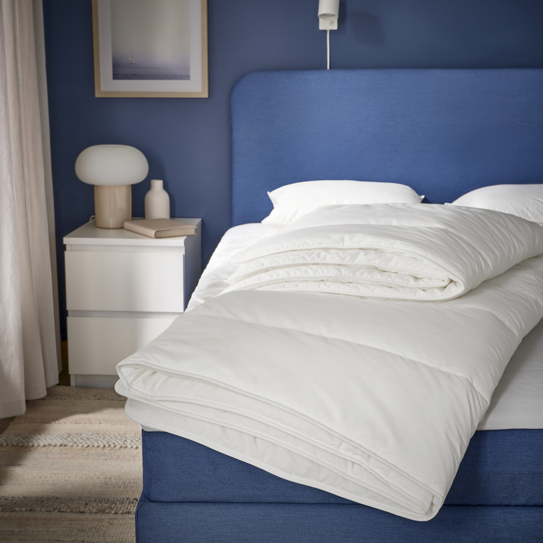 Dormitorio moderno con paredes azul marino y una cama con cabecero tapizado azul, mostrando un edredón blanco doblado sobre la cama y una mesita de noche blanca con lámpara y objetos decorativos.