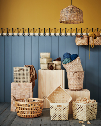 Entryway or room with a two-tone wall (mustard and paneled blue with hooks) and wooden floor, displaying a collection of woven wicker and wooden storage baskets of various sizes and shapes, along with colorful yarns.