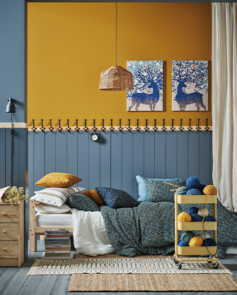 Bedroom with a two-tone wall (mustard and paneled blue with hooks) and a wooden bed with patterned cushions and blue textiles, flanked by a wooden nightstand and a cart with colorful yarns, and decorated with two deer paintings.