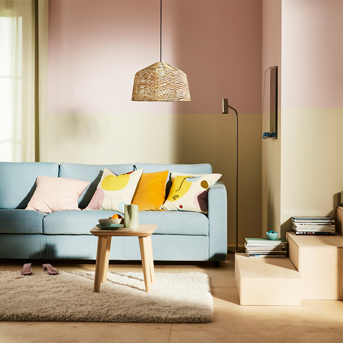 Living room with two-tone walls (pink and beige), a light blue sofa with pastel and yellow decorative cushions, a light wooden coffee table, a fluffy beige rug, and a woven wicker ceiling lamp.