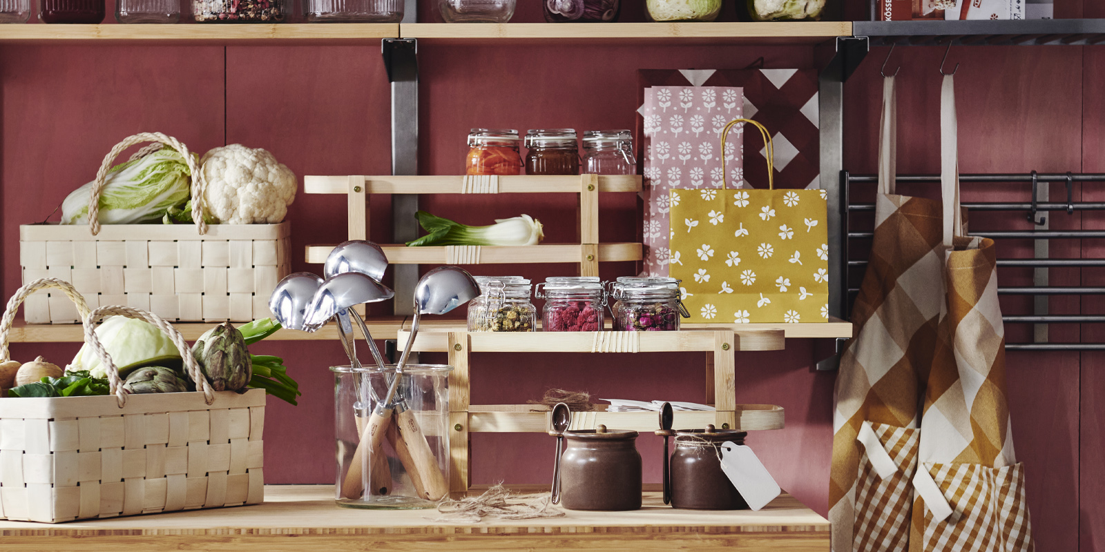 Una cocina con estantes y una mesa adornada con diversos artículos KÖSSEBÄR, como jarras, cestas, paños de cocina, delantales, y más.