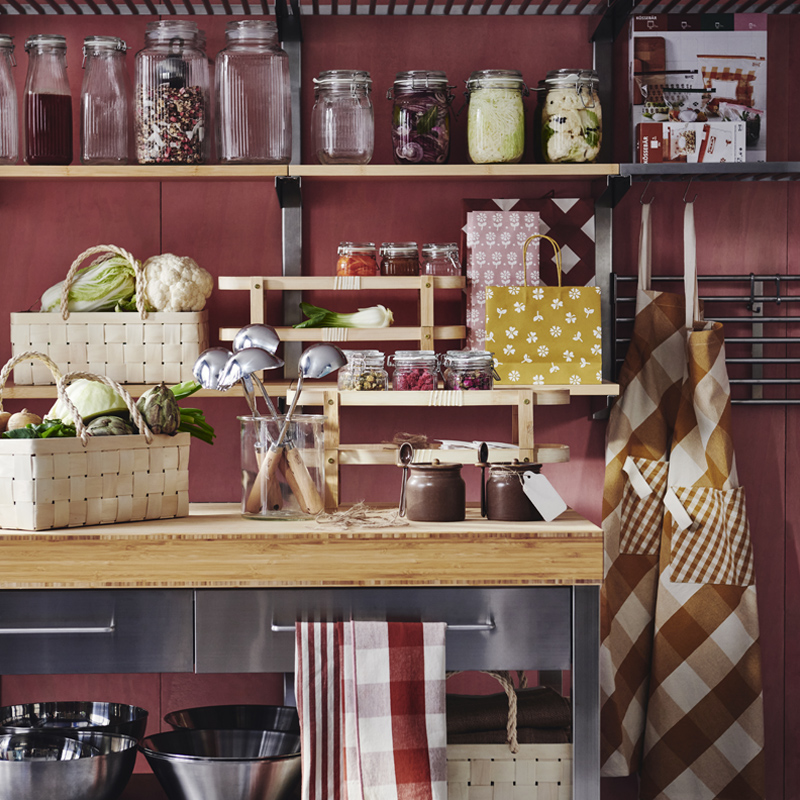 Una cocina con estantes y una mesa adornada con diversos artículos KÖSSEBÄR, como jarras, cestas, paños de cocina, delantales, y más.