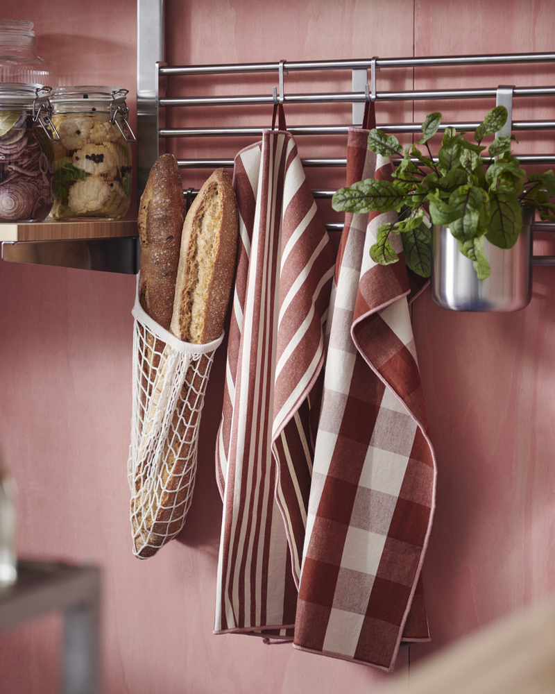 Dos paños de cocina KÖSSEBÄR con estampados variados, una bolsa de malla con pan y una planta en maceta cuelgan de los rieles de una estantería de pared.