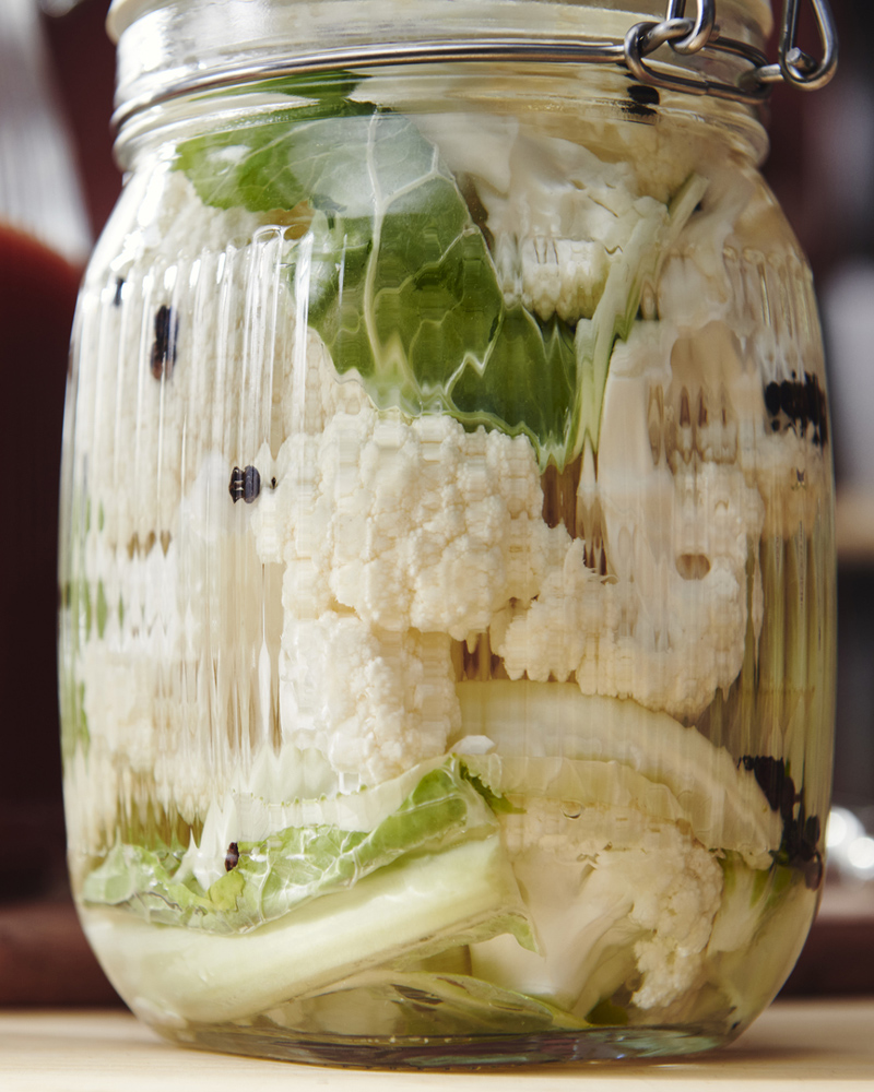 Un tarro de cristal transparente KÖSSEBÄR con tapa, lleno de coliflor y otras verduras, descansa sobre una encimera de madera.