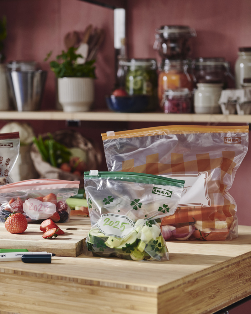 Cuatro bolsas resellables KÖSSEBÄR de diferentes colores, llenas de verduras, bayas y setas, están colocadas sobre una encimera.