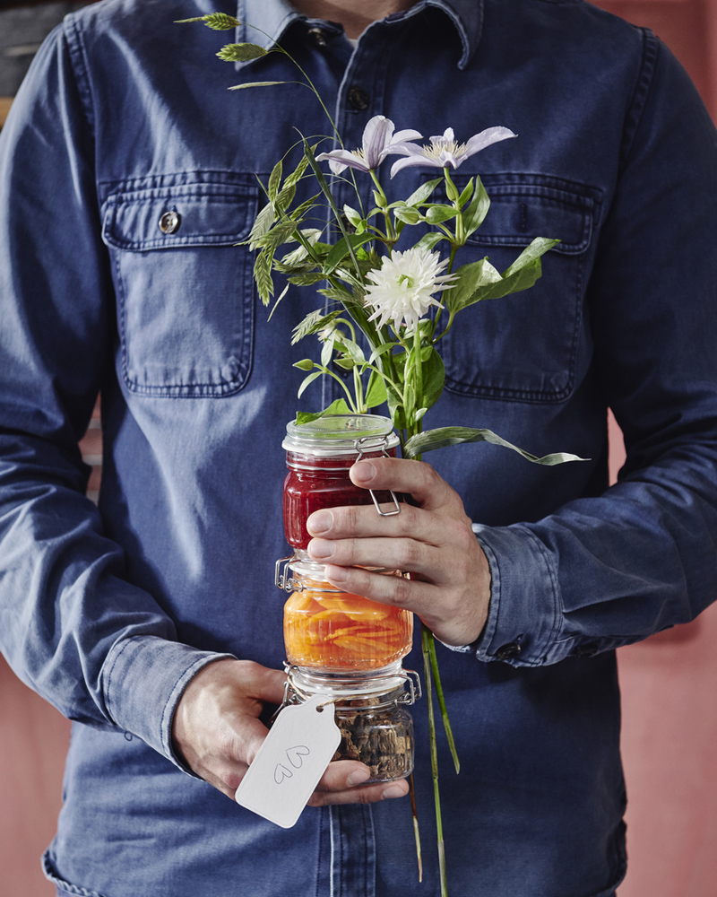 Una persona sostiene unas flores y tres tarros KÖSSEBÄR con tapas llenos de diferentes alimentos delante de sí.