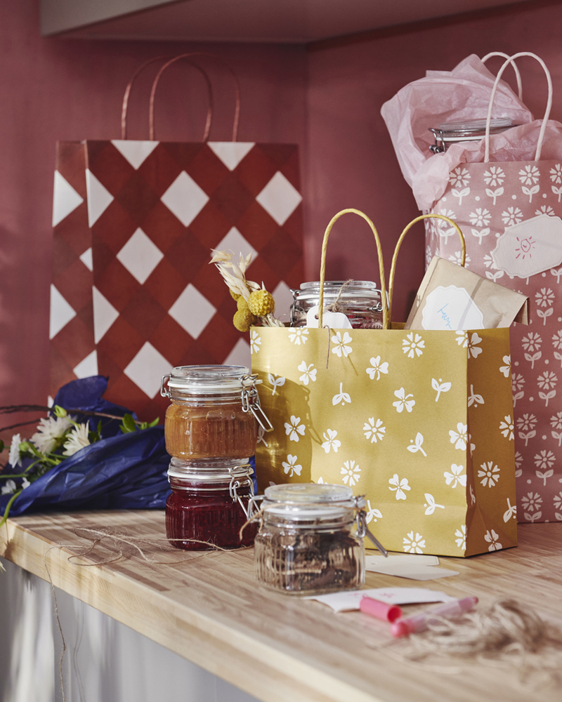 Tres bolsas de regalo KÖSSEBÄR con estampados variados están colocadas sobre una encimera junto a un ramo de flores y varios tarros llenos de alimentos.