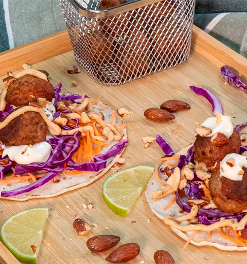 Tacos de albóndigas servidos en un plato de madera, cubiertos con col morada picada, zanahoria rallada, salsa blanca, almendras tostadas y perejil, acompañados de gajos de lima y una cesta metálica con más albóndigas.