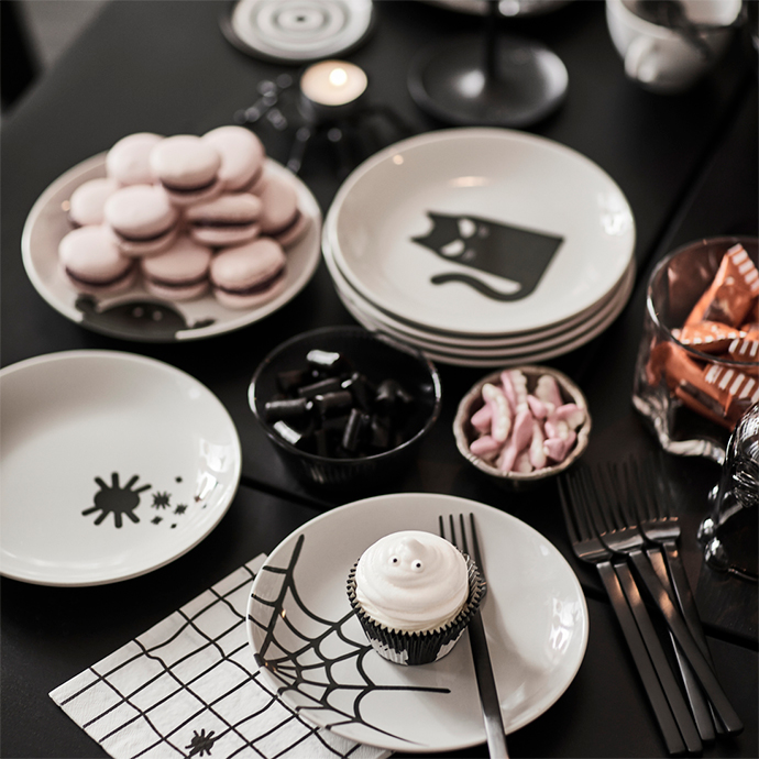 Halloween table decorated with cat and spider-themed plates, spiderweb napkins, trays with pink macarons, black licorice candies, and a white cupcake decorated with eyes, accompanied by black cutlery.