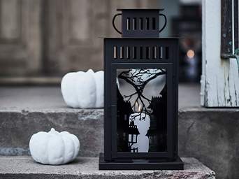 Black decorative lantern with haunted house and bare tree cutouts, illuminated from within by a candle, flanked by two white pumpkins, on outdoor stone steps.