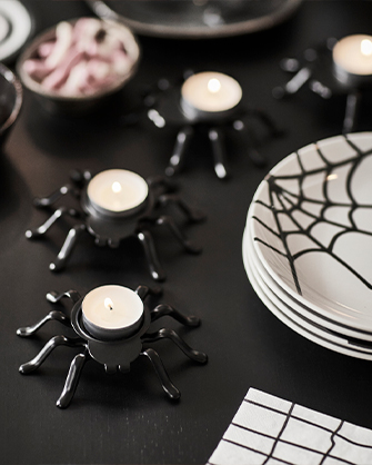 Halloween table decoration with black and white tablecloths and spiderweb-patterned plates, and several black spider-shaped candle holders with lit candles, next to a bowl of sweets.