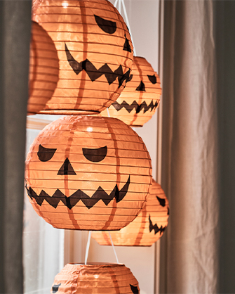 Several orange paper pumpkin-shaped lanterns with illuminated Halloween faces, hanging vertically in front of a window with light curtains, creating a festive and warm ambiance.