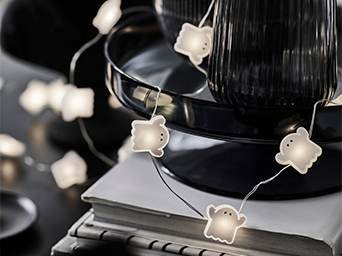 Decorative string lights in the shape of illuminated white ghosts, entwined around a stack of books and a dark vase on a black tray, creating a ghostly and festive ambiance.