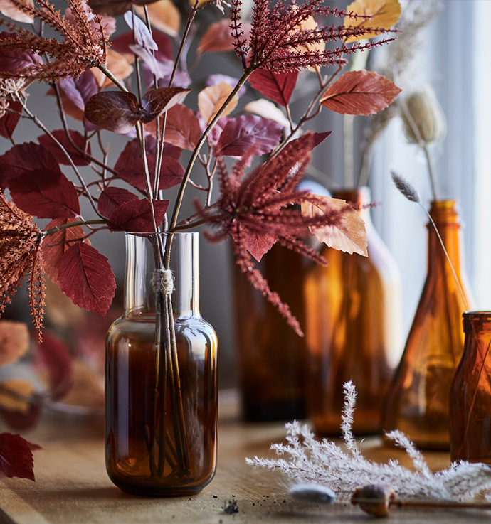 Autumnal arrangement featuring branches with reddish and deep red leaves in an amber-toned glass vase on a wooden surface, accompanied by dark glass bottles and dried grass in the background, creating a warm and natural ambiance.
