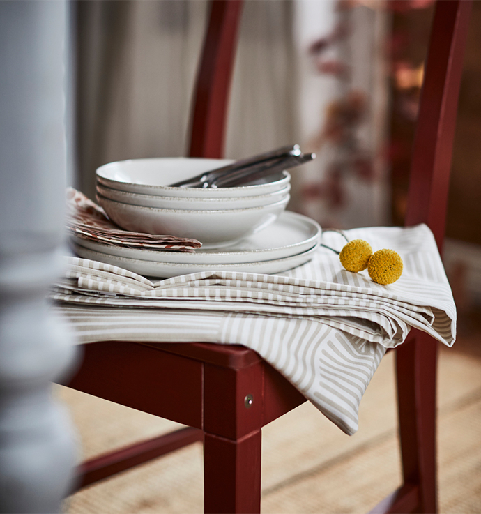 Stack of white plates and bowls with a subtle rim, piled on beige and white striped fabric napkins on the seat of a red wooden chair, with cutlery and two yellow decorative spheres.