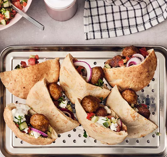 Panes de pita abiertos rellenos con albóndigas, cebolla roja, pepino, tomate y una salsa blanca cremosa, presentados en una bandeja metálica perforada.